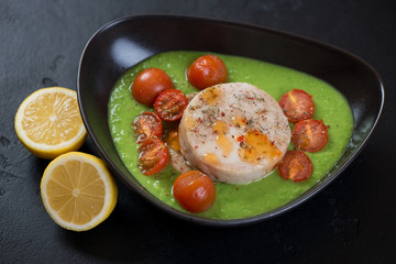 Baked hake medallions with green peas puree and cherry tomatoes in a bowl, studio shot on a black stone background