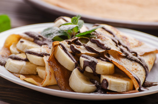 French crepes with chocolate sauce and banana in ceramic dish on wooden kitchen table