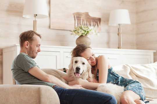 Happy Lovers Newlyweds Enjoying A Weekend In The Room Of Their Cozy Country House Sitting On The Sofa With Their Beloved Dog