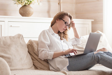 Pensive young woman freelancer working remotely using a laptop while sitting on a sofa in her cozy living room. Concept of comfortable work at home