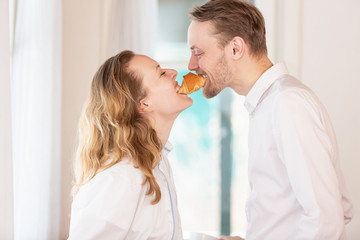 Side view of a funny happy married couple biting a croissant simultaneously from two sides. Concept of breakfast and good relationship