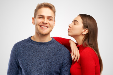 Obraz premium Pleased pretty female, stands sideways, leans at shoulder of her handsome bearded boyfriend, going to kiss in cheek, expresses love and truthful feeling, isolated on white wall in studio