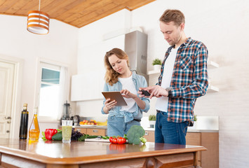Young married couple in casual clothes is looking for a salad recipe that is made from vegetables using a smartphone and tablet while standing in the kitchen of their cozy country house