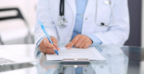 Unknown doctor woman at work, close-up. Female physician filling up medical form while sitting at glass desk at hospital office. Medicine and healthcare concept