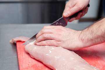 Closeup cook hand cuts pork fillet steak with sharp knife on red plastic cutting board on metal table in restaurant kitchen. Concept steakhouse specializing in grilled meat, live fire