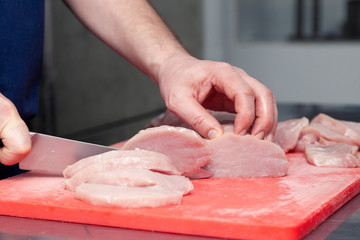 Closeup cook hand cuts pork fillet steak with sharp knife on red plastic cutting board on metal table in restaurant kitchen. Concept steakhouse specializing in grilled meat, live fire