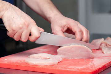 Closeup cook hand cuts pork fillet steak with sharp knife on red plastic cutting board on metal table in restaurant kitchen. Concept steakhouse specializing in grilled meat, live fire