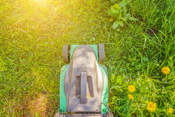 Lawn mower cutting green grass in backyard in sunny day. Gardening country lifestyle background. Beautiful view on fresh green grass lawn in sunlight, garden landscape in spring or summer season