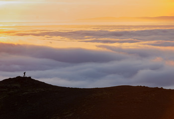 Naklejka na ściany i meble Silhouette of a man on the ridge above the sea of clouds, misty mountains at sunset in Iceland