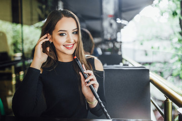 A young beautiful girl smokes hookah on the summer terrace of a modern restaurant. Dressed in a...