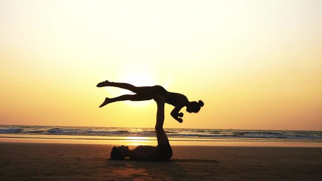 Silhouette of fit sporty couple practicing acrobatic yoga with partner together on the beach in slow motion. Female acrobat is balancing on the legs of her male partner and doing rotation.