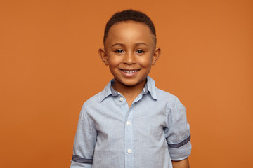 Handsome adorable dark skinned schoolboy wearing neat blue shirt smiling broadly, rejoicing at good mark at school, posing against orange wall background with copy space for your advertising content