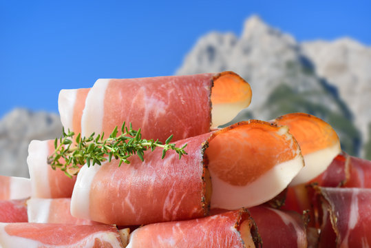 Aufgeschnittener S&uuml;dtiroler Speck vor Bergen in den Alpen &ndash; Sliced South Tyrolean bacon in front of mountains of the alps