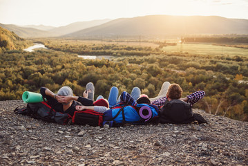 Traveling, tourism and friendship concept. Group of young friends traveling together in mountains. Happy hipster travelers with backpacks lying on the top of mountain at sunset background.