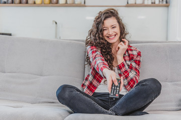Obraz premium A beautiful woman watches the TV and is sitting on the couch and holds the remote control in his hand. A brunette in a shirt looks at the TV with long hair.