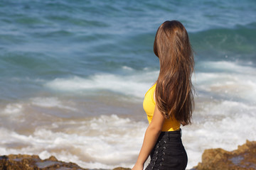 Young dark-haired beautiful European girl in yellow swimsuit smiling and standing on the background of the sea and splashes on a Sunny day
