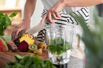 Vegan woman cutting pineapple on wooden board to prepare healthy fruit smoothie with kiwi and beetroot