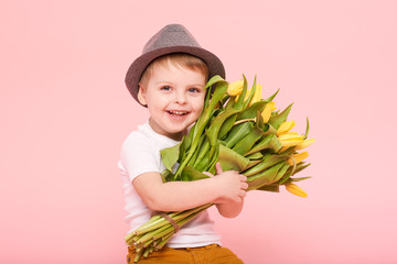 Adorable smiling child with spring flower bouquet looking at camera isolated on pink. Little toddler boy in hat holding yellow tulips as gift for mom