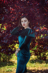 Portrait of beautiful young brunette woman with bright makeup, wearing emerald green velvet dress, standing  in a park, near the tree with red leaves