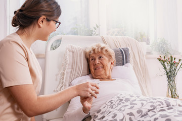 Positive senior woman lying in bed, helpful doctor in beige uniform supporting her