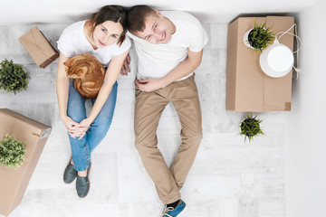 A young married couple with a red cat, a man and a woman, are sitting on the floor in a bright room against the background of cardboard boxes, green plants and things.