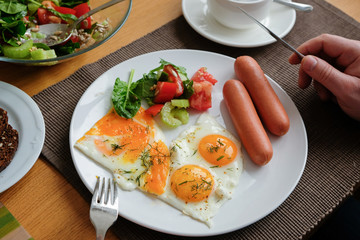 Somebody eating breakfast. Knife and fork in hand. American style breakfast with fried eggs, sausage, salad and toast.