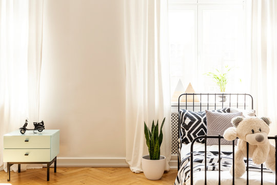 Teddy bear on child's bed in white bedroom interior with plant and copy space on the wall. Real photo