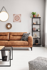Leather sofa with black and white pillow in real photo of sitting room interior with metal rack with books, decor and fresh plant and mirror, clock and poster on the wall