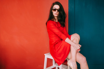 Beautiful fashion girl with dark long hair, spanish appearance in sunglasses and red elegnat dress posing on blue red wall in studio. Indoor lifestyle portrait of stylish woman. Aamzing brunette babe