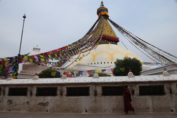 A monk passing by and spinning prayer wheels at Boudhanath stupa on a cloudy day. Kathmandu, Nepal.