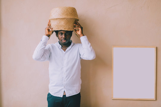 Closeup lifestyle portrait of strange young african black man holding straw basket above his head on textured wall background and looking at camera. Dark-skinned nigerian boy  indoor funny portrait.
