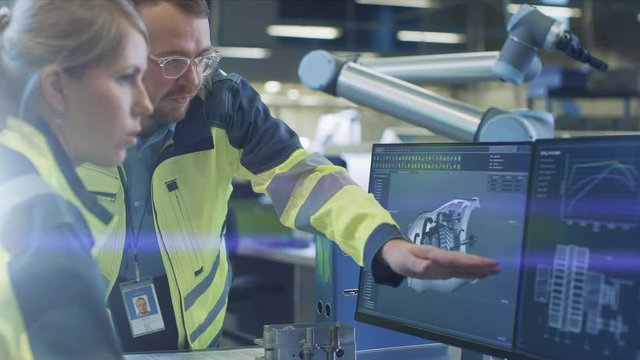 At the Factory: Male Mechanical Engineer and Female Chief Engineer Discuss Operational 3D CAD Designed Engine Prototype Shown on Screen. In Background Futuristic factory with Automated Robotic Hands