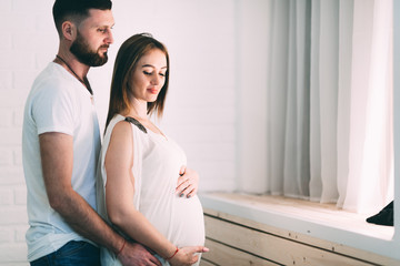 Fototapeta premium Young pregnant couple near the window of the house, on the background of a brick white wall and a wooden window sill