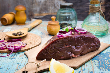 A piece of raw beef liver on a cutting board, onion, lemon and spices for cooking on a wooden table