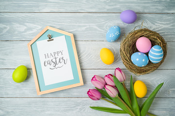 Easter holiday background with easter eggs in bird nest, note and tulip flowers on wooden table.