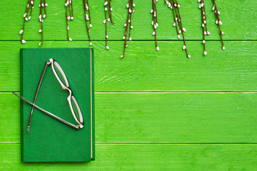 A book in a hard green cover on a green wooden background, glasses and willow branches. Top view. Copy space