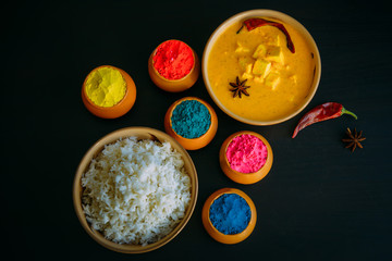 Holi indian festival of colors. Food with colours, steam rice, panir butter masala, pepper chilie, starnise. Powder colours arranged over black background. Selective focus