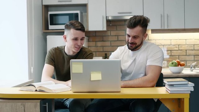 Two guys learn and work on a computer on project using a textbook sitting in the kitchen at home. Talking each other.