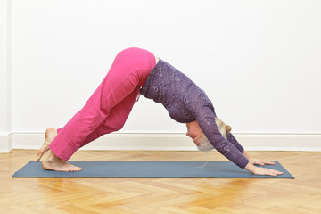 Senior woman exercising yoga at home on a mat in front of a white wall, asana walking dog