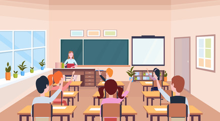 pupils raising hands to answer question teacher sitting desk education concept modern school classroom interior chalk board horizontal flat