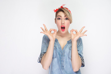 Portrait of surprised beautiful young woman in casual blue denim shirt with makeup and red headband standing with ok sign and looking at camera. indoor studio shot, isolated on white background.