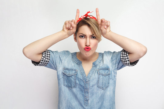 Portrait of funny beautiful young woman in casual blue denim shirt with makeup and red headband standing with horns hands on head and looking at camera. indoor studio shot isolated on white background