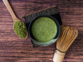 A cup of green tea latte and matcha powder in spoon with bamboo whisk on wooden background.