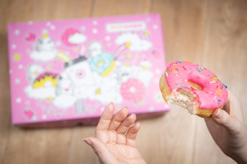 The donut with pink icing in the hand of the girl on the background of a pink box and a wooden background