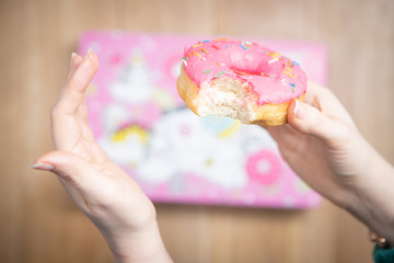 The donut with pink icing in the hand of the girl on the background of a pink box and a wooden background