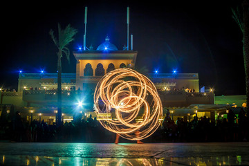 Night performance fire show in front of a crowd of people on the street in Egypt, palace and palm trees in the soft focus background.