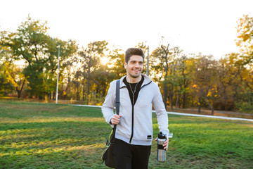 Sportsman outdoors in park listening music with earphones walking with bag.