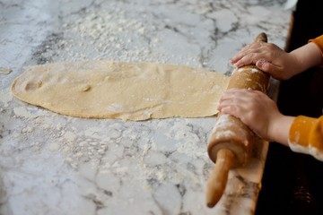 Child's hands rolling out dough with wooden rolling pin and some flour on white marble surface