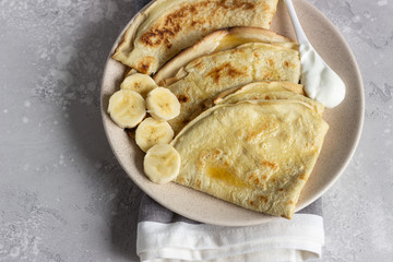 Thin pancakes or crepes with natural yogurt, honey and banana on a plate. Light concrete background. Copy space.