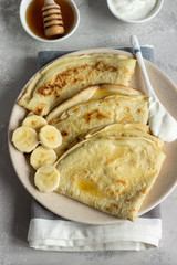 Thin pancakes or crepes with natural yogurt, honey and banana on a plate. Light concrete background. Copy space.